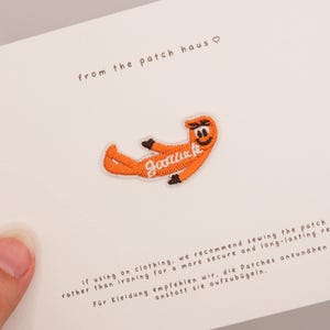 Puede incluir: Un parche bordado de color naranja que muestra un personaje de dibujos animados en posición reclinada con el texto cursivo blanco "goodluck" en el centro. El parche se presenta sobre una tarjeta blanca con el texto "from the patch haus" e instrucciones.