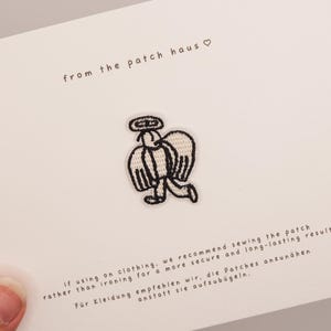 Könnte beinhalten: Ein kleiner gestickter Aufnäher mit einer schwarz-weißen Strichzeichnung eines Engels mit Flügeln und Heiligenschein. Der Aufnäher ist auf einer weißen Karte zentriert, die den Text from the patch haus und eine Nähanleitung enthält.