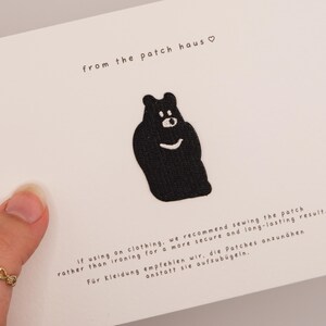 May include: A black embroidered bear patch centred on a white card. The card features the text from the patch haus with a heart symbol, and instructions in English and German regarding sewing the patch onto clothing for a secure result.