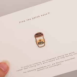 Puede incluir: Un parche bordado con forma de taza de café con tapa marrón y cuerpo color canela que presenta el texto COFFEE en hilo negro. El parche está sujeto a una tarjeta blanca con el texto from the patch haus e instrucciones de aplicación.