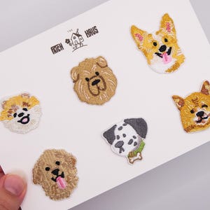 Op de afbeelding: Zes geborduurde hondenpatches gerangschikt op een witte kaart met de tekst THE PATCH HAUS. De patches tonen een Corgi, een Shar-Pei, een Pomeriaan, een Dalmatiër, een Shiba Inu en een Golden Retriever, allemaal uitgevoerd in gedetailleerde draadkleuren.