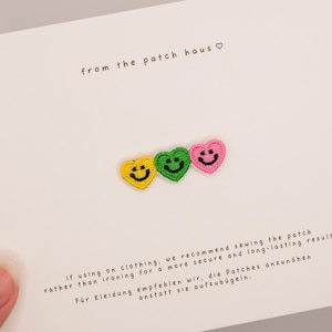 May include: Three small embroidered heart-shaped patches in yellow, green, and pink, each featuring a black stitched smiley face. The patches are attached to a white card with the text from the patch haus and sewing instructions.