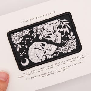 May include: A rectangular embroidered patch featuring a black background with white thread detailing two skulls, roses, leaves, a crescent moon, and small stars. The text "from the patch haus" is printed above the patch on a white backing card.