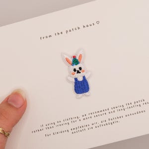 May include: An embroidered patch of a white rabbit wearing a blue overall and a colorful party hat, centered on a white card with the text from the patch haus and instructions for sewing the patch onto clothing for a secure result.