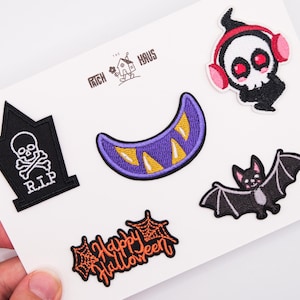 Könnte beinhalten: Ein Set aus fünf bestickten Aufnähern mit einem schwarzen Grabstein mit weißem Totenkopf, einer violetten Mondsichel mit gelben Zähnen, einem Geist mit rosa Kopfhörern, einer schwarzen Fledermaus und dem orangefarbenen Text Happy Halloween.