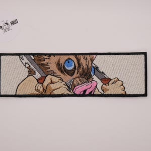 Op de afbeelding: Een rechthoekige geborduurde patch met een bruin zwijnachtig personage met helderblauwe ogen en een roze snuit, die twee gekartelde messen vasthoudt. Het ontwerp staat op een lichtgrijze achtergrond met een dikke zwarte rand en afwerking.