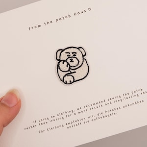 Puede incluir: Un parche bordado blanco que muestra un oso de dibujos animados con contorno negro sentado con las patas recogidas. El parche está sujeto a una tarjeta blanca con el texto from the patch haus e instrucciones en inglés y alemán.