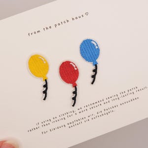 Può includere: Tre toppe ricamate a forma di palloncino in giallo, rosso e blu, ciascuna con un filo nero. Il cartoncino bianco di presentazione riporta la scritta from the patch haus e le istruzioni che consigliano di cucire le toppe sui vestiti.