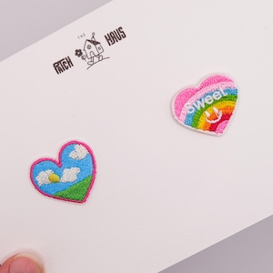 以下が含まれることがあります： THE PATCH HAUSという文字が書かれた白い台紙に、ハート型の刺繍ワッペンが2つ付いています。1つは青い空、白い雲、黄色い太陽、緑の草が描かれています。もう1つは虹色のハートで、スマイリーフェイスとsweetという文字が刺繍されています。