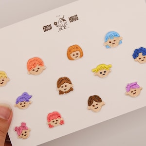以下が含まれることがあります： 黄色、オレンジ、赤、紫、茶色、青、ピンクなど、さまざまな髪色の漫画の顔が刺繍された12個の小さなワッペン。白い台紙には「THE PATCH HAUS」という文字が印刷されており、ワッペンがその上に配置されています。
