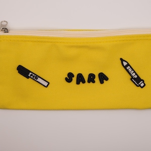 Puede incluir: Un estuche rectangular de color amarillo brillante con cierre de cremallera y un ojal metálico en el lado izquierdo. El frente presenta parches bordados en negro de un bolígrafo, el nombre SARA en letras negritas y un marcador con la palabra SHARP.
