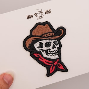 May include: An embroidered patch featuring a white skull wearing a brown cowboy hat and a red bandana. The patch has a black border and is displayed on a white card with the text THE PATCH HAUS and a small house illustration printed at the top.