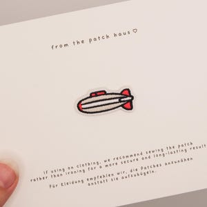 Könnte beinhalten: Ein gestickter Aufnäher in Form eines Zeppelins mit hellgrauem Körper, schwarzen Umrandungen und roten Akzenten. Der Aufnäher ist auf einer weißen Karte befestigt, die den Text from the patch haus und Anweisungen zum Annähen enthält.