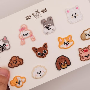 May include: A collection of twelve embroidered dog patches arranged on a white card labeled THE PATCH HAUS. The patches feature various dog breeds in colors including white, brown, grey, and orange, with some dogs wearing pink bows or accessories.