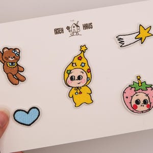 May include: A set of five embroidered patches on a white card labelled THE PATCH HAUS. The designs include a brown teddy bear, a yellow star-patterned hooded figure, a pink strawberry-themed character, a blue heart, and a yellow shooting star.
