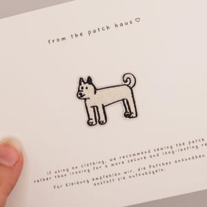 May include: A white embroidered patch featuring a black-outlined dog with a curled tail, centred on a white card. The card displays the text from the patch haus and instructions recommending sewing the patch onto clothing for durability.