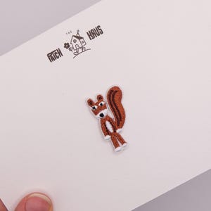 May include: An embroidered iron-on patch featuring a brown squirrel with a white chest and face, standing upright with a curled tail. The patch is attached to a white backing card printed with the text THE PATCH HAUS and a small house icon.