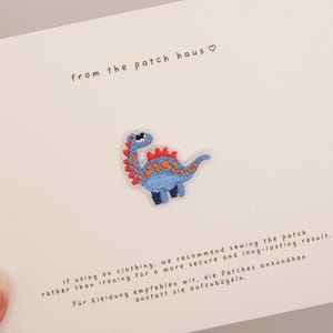 May include: An embroidered dinosaur patch featuring a light blue body, dark blue legs, and red spikes along the back with orange spots. The patch is centred on a white card with the text from the patch haus and instructions for application.
