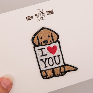 Può includere: Una toppa ricamata raffigurante un cucciolo color marrone chiaro che tiene un cartello bianco con la scritta I LOVE YOU e un cuore rosso. La toppa ha un bordo nero ed è esposta su un cartoncino bianco con la scritta THE PATCH HAUS e una casa.