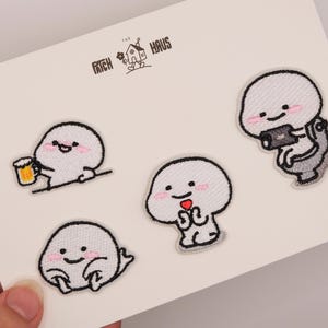 May include: Four embroidered patches on a white card featuring the text THE PATCH HAUS. The patches depict white, round-headed characters: one holding a beer mug, one sitting on a toilet with a phone, one holding a red heart, and one sitting down.