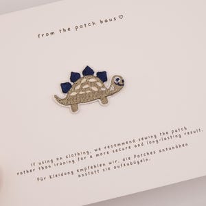 Puede incluir: Un parche bordado de estegosaurio en beige y azul oscuro, centrado en una tarjeta blanca. La tarjeta incluye el texto 'from the patch haus' con un símbolo de corazón e instrucciones que recomiendan coser el parche a la ropa.
