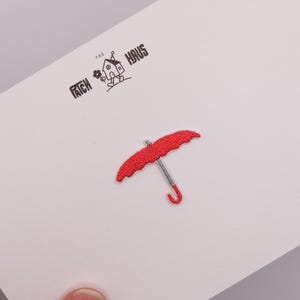 May include: A red umbrella embroidered patch with a grey handle and shaft, centered on a white card. The card features the black printed text THE PATCH HAUS with a small graphic of a house with legs holding a flower above the umbrella patch.