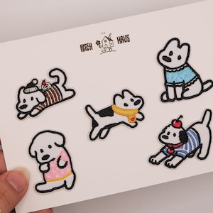 May include: A set of five embroidered dog patches on a white card labeled PATCH HAUS. The patches feature white dogs in various outfits: a brown sweater with a beanie, a blue sweater, a yellow scarf, a pink shirt, and a blue striped shirt with a cherry.