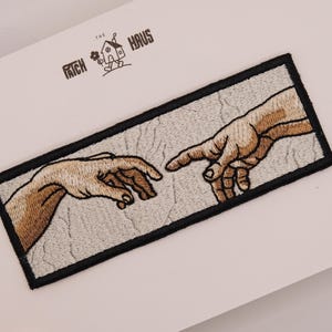 May include: A rectangular embroidered patch featuring a recreation of the Creation of Adam hands reaching toward each other. The design uses tan and brown thread for the skin tones against a textured light gray background with a black border.
