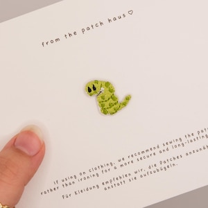 May include: A small, light green embroidered patch shaped like a worm with dark eyes, centred on a white display card. The card features the text from the patch haus and instructions in English and German regarding sewing the patch onto clothing.