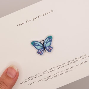 Op de afbeelding: Een geborduurde vlinderpatch in tinten blauw, turquoise en paars met zwarte voelsprieten. De patch is gecentreerd op een witte kaart met de tekst from the patch haus en instructies in het Engels en Duits over het aanbrengen ervan.
