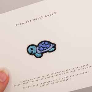 Puede incluir: Un parche bordado de tortuga con cabeza y extremidades de color azul claro y un caparazón con patrones en azul oscuro y morado. El parche está sujeto a una tarjeta blanca impresa con el texto from the patch haus e instrucciones de costura.