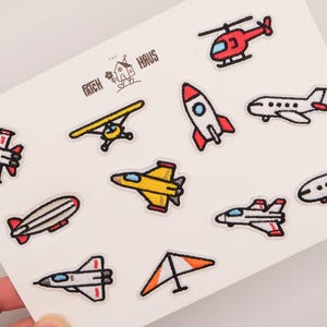 May include: A collection of eleven embroidered patches featuring various aircraft including a red helicopter, a white and red rocket, a yellow jet, a hang glider, a blimp, and several aeroplanes. The text THE PATCH HAUS is printed in black.