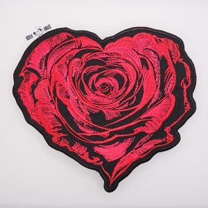 Puede incluir: Un parche bordado en forma de corazón con un diseño detallado de una rosa roja sobre un fondo negro. El bordado utiliza hilo rojo vibrante para formar los pétalos de la rosa, que tiene forma de corazón. El texto PATCH HAUS es visible.