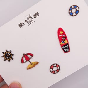 Könnte beinhalten: Ein Set aus fünf bestickten Aufnähern mit Strandmotiven, darunter ein Steuerrad, ein rot-weißer Sonnenschirm, ein rotes Surfbrett mit dem Text Sunny Day sowie zwei Rettungsringe in Rot und Blau, die auf einer weißen Karte präsentiert werden.