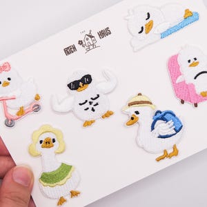 Puede incluir: Un conjunto de seis parches bordados termoadhesivos con patos blancos en diversas actividades, incluyendo uno en un patinete rosa, otro con gafas de sol, uno sentado en una silla rosa y otro con sombrero y mochila. Se lee el texto THE PATCH HAUS.