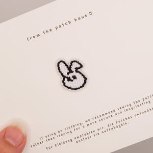 Pode incluir: Um pequeno emblema bordado branco em forma de cabeça de coelho com contornos pretos. O emblema está fixado num cartão branco com o texto from the patch haus e instruções para coser o emblema na roupa para uma maior durabilidade.