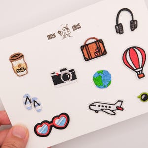 Peut inclure: Une collection d'écussons brodés sur le thème du voyage sur une carte blanche, comprenant une tasse de café, une valise, un casque, un appareil photo, un globe, une montgolfière, des tongs, un avion, des lunettes et des lunettes en forme de cœur.