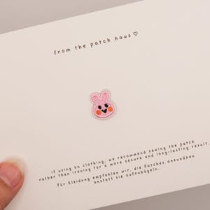 Könnte beinhalten: Ein kleiner, rosa gestickter Hasenaufnäher mit schwarzen Augen, einer schwarzen Nase und orangen Wangen, zentriert auf einer weißen Karte. Die Karte enthält den Text 'from the patch haus' und Anweisungen zum Annähen des Aufnähers.