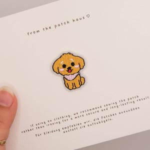 Puede incluir: Un parche bordado que muestra un cachorro de color marrón dorado con un pañuelo a cuadros rosas y blancos. El parche está sujeto a una tarjeta blanca con el texto from the patch haus e instrucciones que recomiendan coserlo a la ropa.