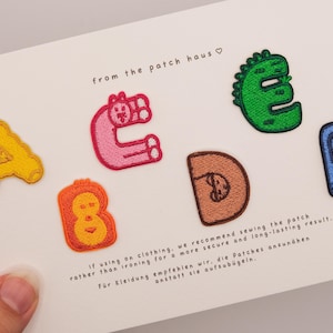 May include: A set of six embroidered letter patches arranged on a white card. The letters A, B, C, D, E, and F are featured in yellow, orange, pink, brown, green, and blue. The card includes the text from the patch haus and sewing instructions.