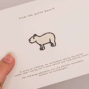 May include: A white embroidered capybara patch centered on a rectangular card. The card features the text from the patch haus with a heart symbol, and instructions in English and German regarding sewing the patch onto clothing for durability.