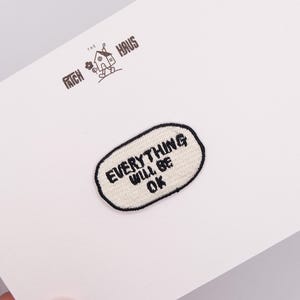 Op de afbeelding: Een ovaalvormige geborduurde patch met een crèmekleurige achtergrond en een dikke zwarte rand. De tekst "EVERYTHING WILL BE OK" is in zwarte hoofdletters in het midden geborduurd. De patch is bevestigd op een witte kaart met het logo "THE PATCH HAUS".