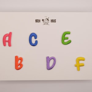 Op de afbeelding: Een set van zes geborduurde alfabet-patches op een witte kaart met het label THE PATCH HAUS. De patches bestaan uit een roze hoofdletter A, een blauwe kleine letter c, een groene hoofdletter E, een oranje kleine letter b, een paarse kleine letter d en een gele kleine letter f.