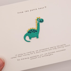 May include: An embroidered dinosaur patch in shades of green with yellow spikes and a white belly, centered on a white card. The card features the text from the patch haus and instructions for sewing the patch onto clothing in English and German.