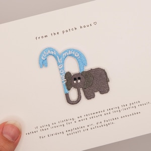 May include: An embroidered patch featuring a grey elephant holding a light blue candy cane with the text "from the patch haus" above it. The card includes instructions in English and German regarding sewing the patch onto clothing.