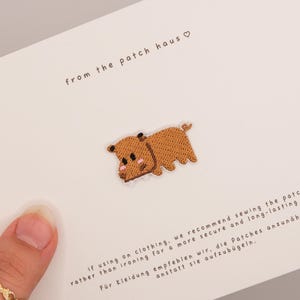 May include: A small, brown, embroidered dog patch featuring a cartoon-style design with dark brown ears, a pink nose, and a small tail. The patch is attached to a white card with the text from the patch haus and sewing instructions.