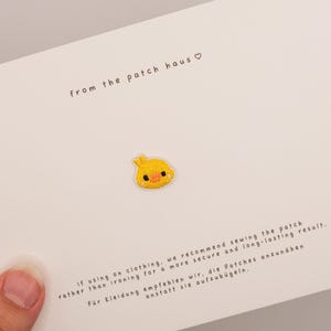 Könnte beinhalten: Ein kleiner gelber gestickter Küken-Aufnäher, zentriert auf einer weißen Karte. Die Karte enthält den Text from the patch haus mit einem Herzsymbol sowie Anweisungen, den Aufnäher für ein sicheres und langlebiges Ergebnis anzunähen.