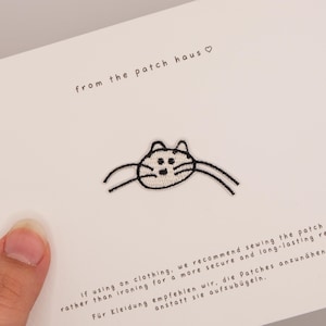 Op de afbeelding: Een witte kaart met de tekst from the patch haus met een hartsymbool en een geborduurde kattenkop-patch in het midden. De kaart bevat instructies in het Engels en Duits over het naaien van de patch op kleding voor een duurzaam resultaat.