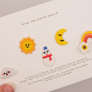 Puede incluir: Un conjunto de cinco parches bordados sobre una tarjeta blanca que incluye un sol sonriente, un muñeco de nieve con bufanda roja, una luna creciente, un arcoíris con flores en los extremos y una pequeña nube. La tarjeta incluye el texto from the patch haus.