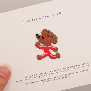Op de afbeelding: Een geborduurde patch met een bruine beer in een rood trainingspak en hoofdband, afgebeeld in een hardloophouding. De patch is bevestigd op een witte kaart met de tekst "from the patch haus" en instructies voor het naaien van de patch op kleding.